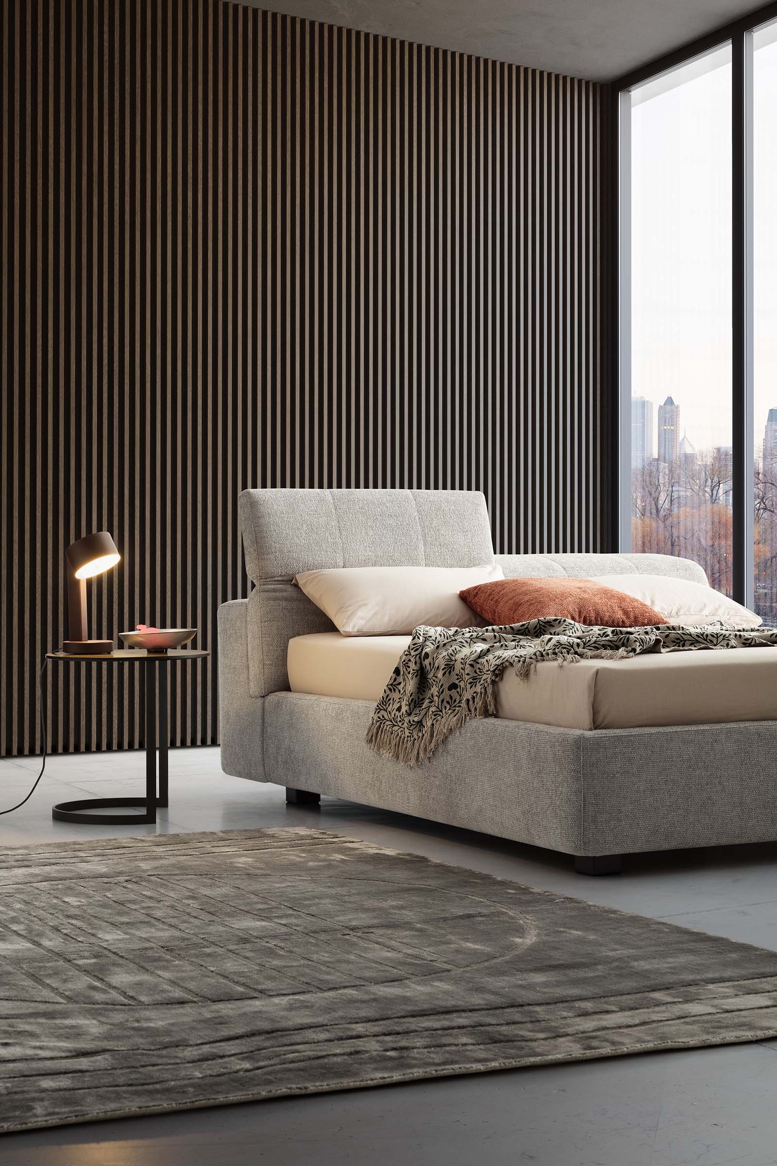 Stanza di città arredata con letto moderno grigio abbinato a tavolino e lampada moderni